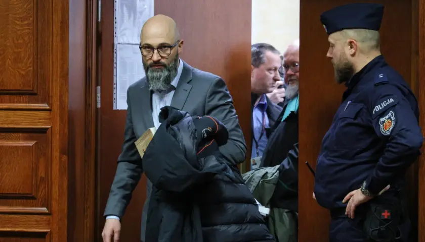 Mężczyzna w garniturze z książką i kurtką, obok policjant i inni mężczyźni w pomieszczeniu.
