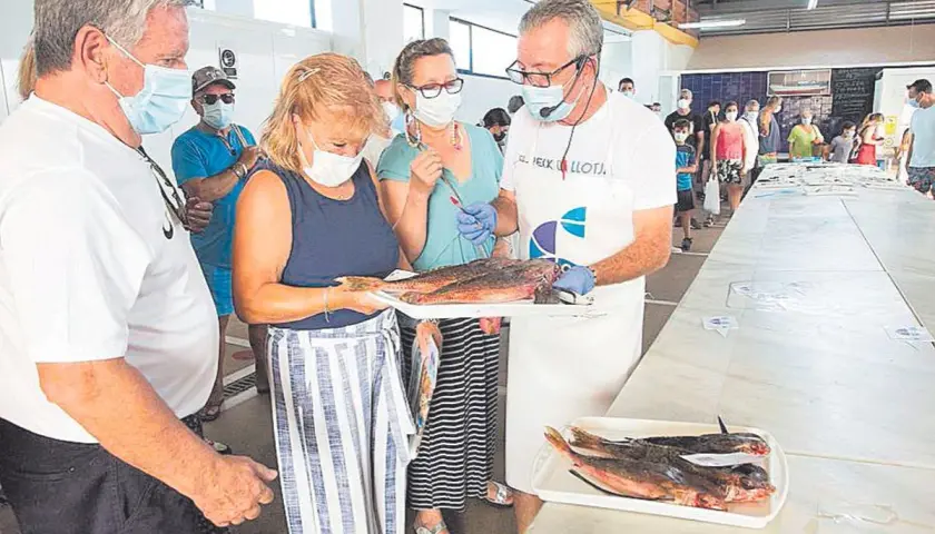 En la lonja del pescado, un hombre con delantal muestra unos peces a una mujer. Otras personas observan.