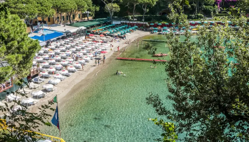Plage animée d'une ville de la Riviera italienne, avec des parasols blancs, des chaises longues et des baigneurs dans l'eau cristalline.
