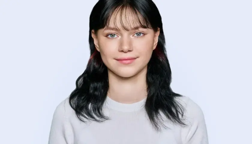 Portret młodej kobiety z czarnymi włosami i grzywką, w białej bluzce, na jasnym tle.