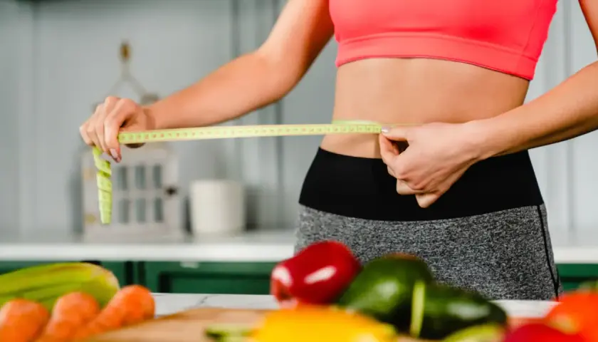 Mujer mide su cintura con cinta métrica, rodeada de verduras frescas. ¿La menopausia adelgaza o engorda?