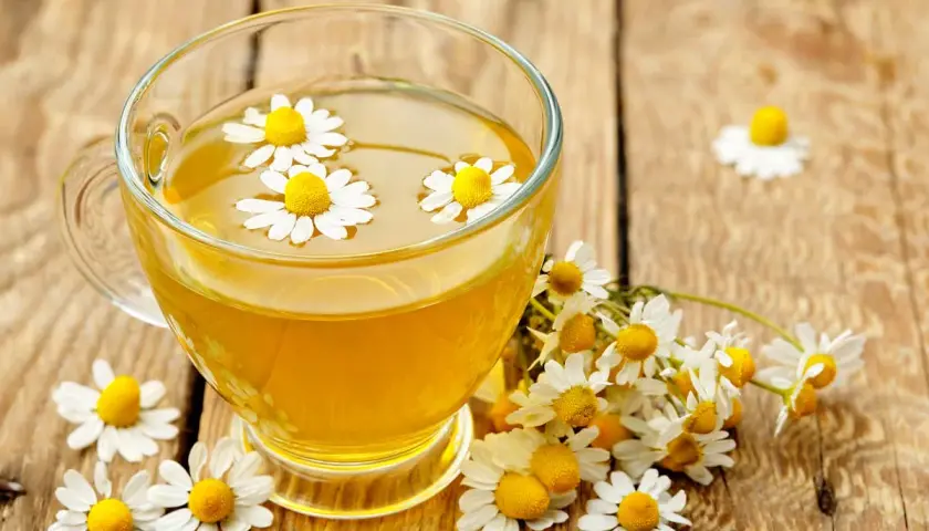 Une tasse de tisane dorée avec des fleurs de camomille flottantes, entourée de fleurs fraîches. Un rappel des bienfaits apaisants de la camomille.