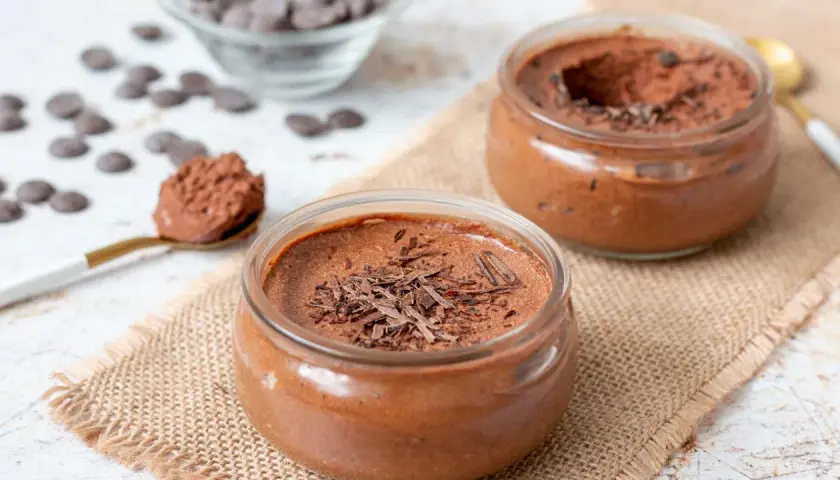 Deux pots de mousse au chocolat, l'un garni de copeaux de chocolat noir, sur une toile de jute. Une cuillère dorée présente une portion de cette délicieuse mousse.