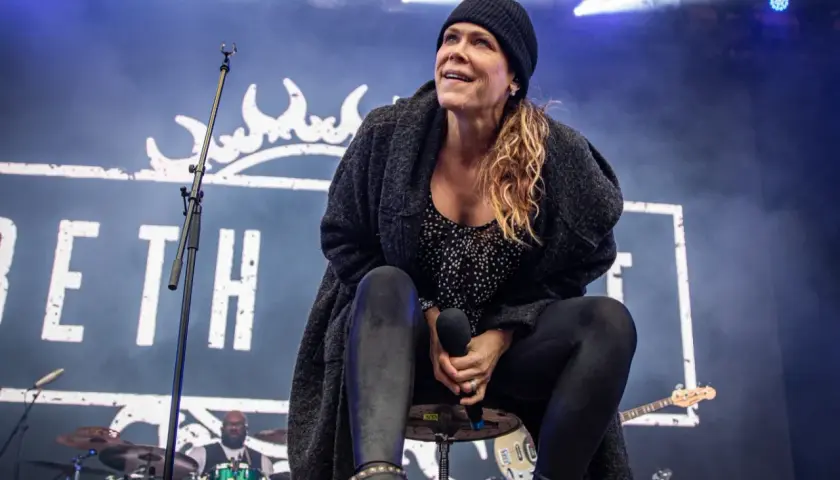 Beth Hart na scenie, w czapce i swetrze, śpiewa swoje utwory. W tle logo z koroną i perkusista.