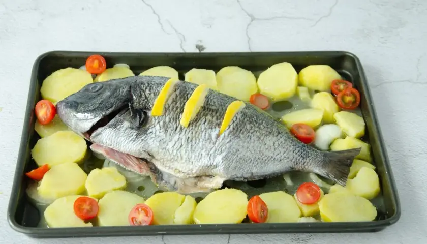 Un sargo pez entero, listo para hornear, rodeado de rodajas de patata y tomates cherry en una bandeja.