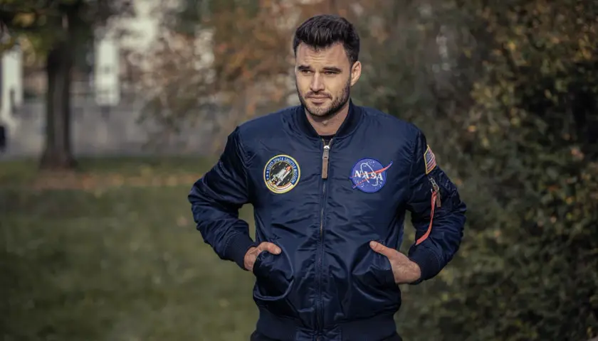 Mężczyzna w granatowej kurtce bomberce Alpha Industries z naszywkami NASA i misji wahadłowca. Alpha Industries to firma znana z odzieży inspirowanej lotnictwem.