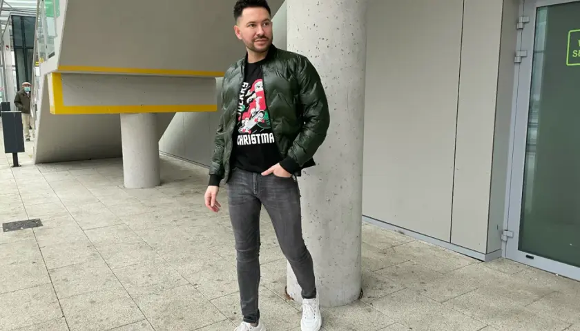 Mężczyzna w zielonej bomberce i szarych jeansach, zastanawia się, jakie buty do bomberki będą najlepsze. Postawił na białe sneakersy.