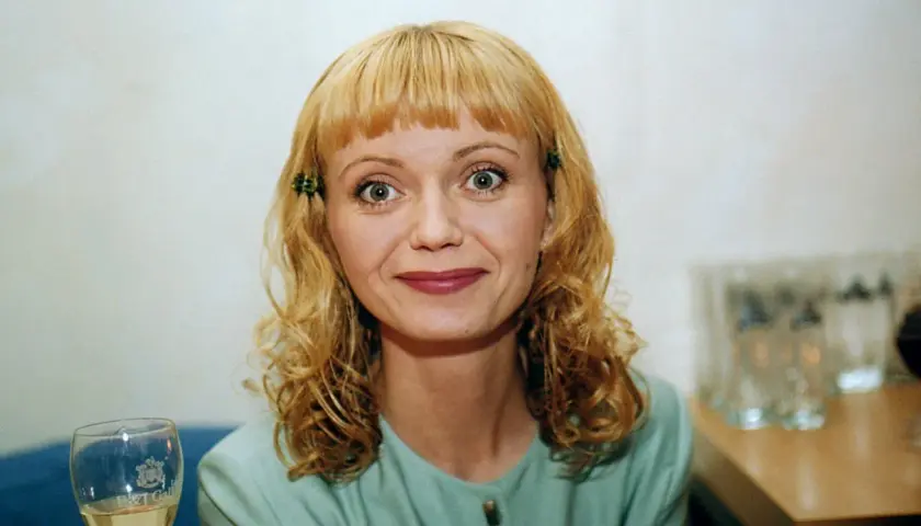Olga Borys z blond włosami i zielonymi spinkami, w zielonej bluzce, z kieliszkiem wina.