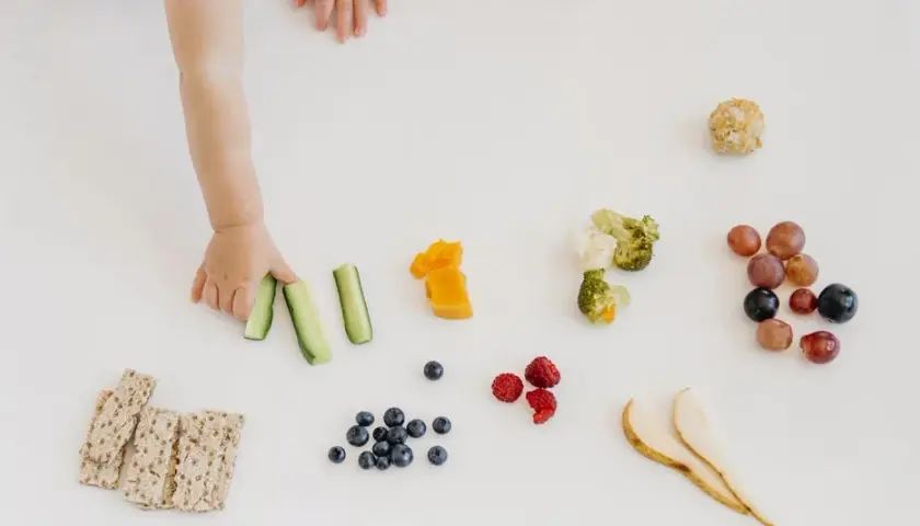 Mano di bimbo afferra bastoncini di cetriolo, parte di un pasto con tagli sicuri svezzamento: frutta, verdura e cracker.