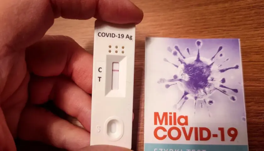 Ręka trzyma test antygenowy COVID-19 z jedną kreską. Obok opakowanie z grafiką wirusa i napisem "Szybki test antygenowy".