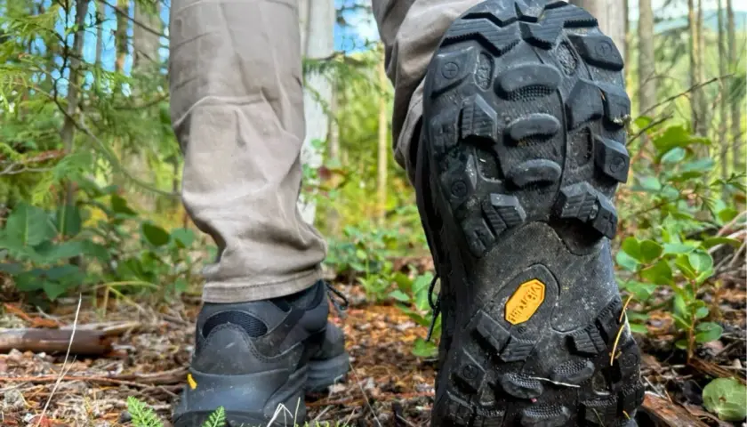 Close-up of a person's hiking boots on a forest trail, showcasing the aggressive tread pattern and the iconic yellow Vibram sole logo, highlighting the durability and grip of various Vibram sole types.
