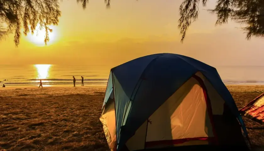 Wschód słońca nad morzem, gdzie rozbito luksusowe pole namiotowe. Ludzie spacerują po plaży, a namioty kuszą komfortem.