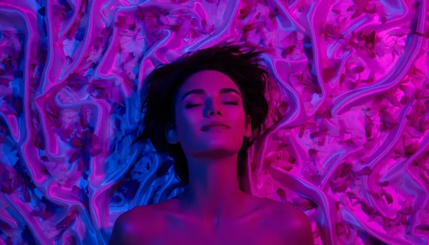 Donna con occhi chiusi, immersa in un letto di seta e petali, illuminata da luci blu e rosa. Un'immagine che evoca il piacere e il mistero del perché godiamo quando veniamo.