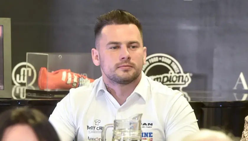 Grzegorz Collins, młody mężczyzna w białej koszuli, z tatuażem na szyi, siedzi przy stole. W tle widoczny pomarańczowy but sportowy.