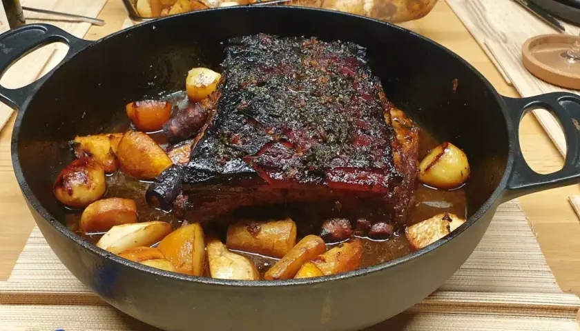 Poitrine de porc au four cuisson lente, dorée et savoureuse, accompagnée de pommes de terre rôties dans un plat en fonte.