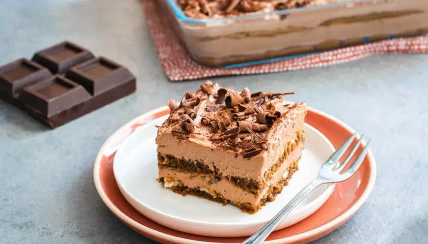 Una fetta di tiramisù cioccolato, ricca e cremosa, guarnita con riccioli di cioccolato. Accanto, pezzi di cioccolato fondente e un'intera teglia.