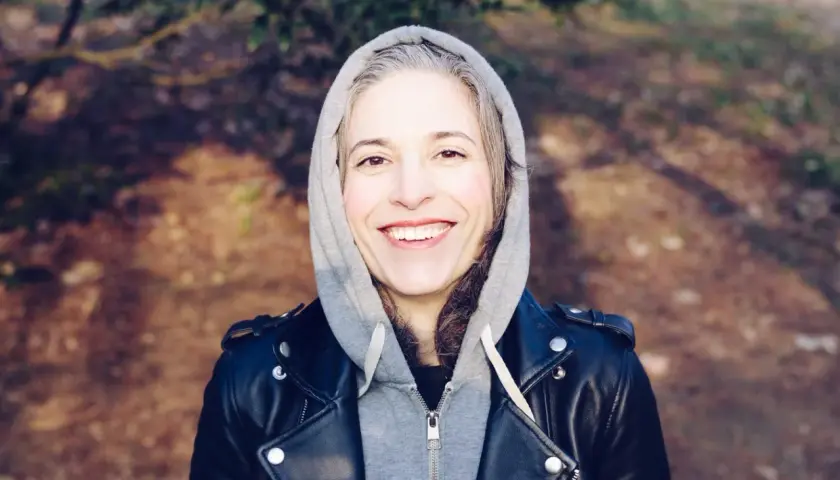 Uśmiechnięta kobieta w szarej bluzie z kapturem i czarnej ramonesce. Jej włosy mają naturalny kolor, z widocznymi siwymi odrostami.