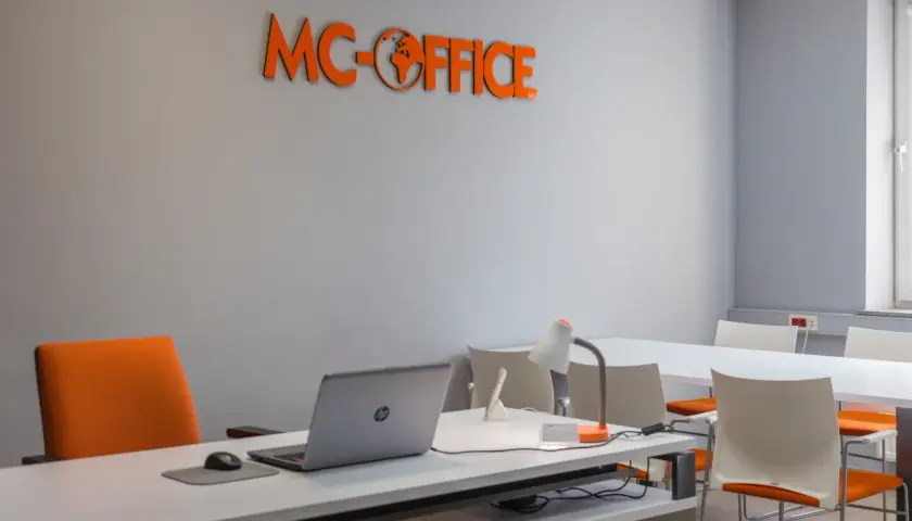 Pomarańczowe logo "MC-OFFICE" na szarej ścianie nad biurkiem z laptopem i krzesłem.