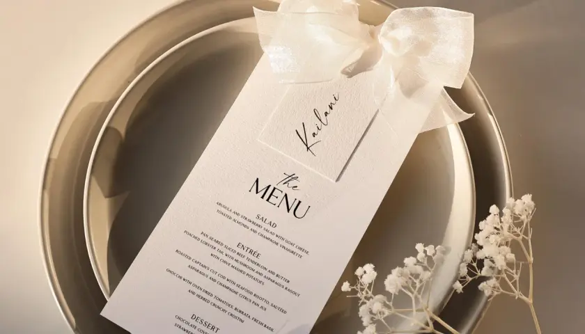 Menu de mariage élégant sur une assiette, orné d'un ruban et de petites fleurs blanches.