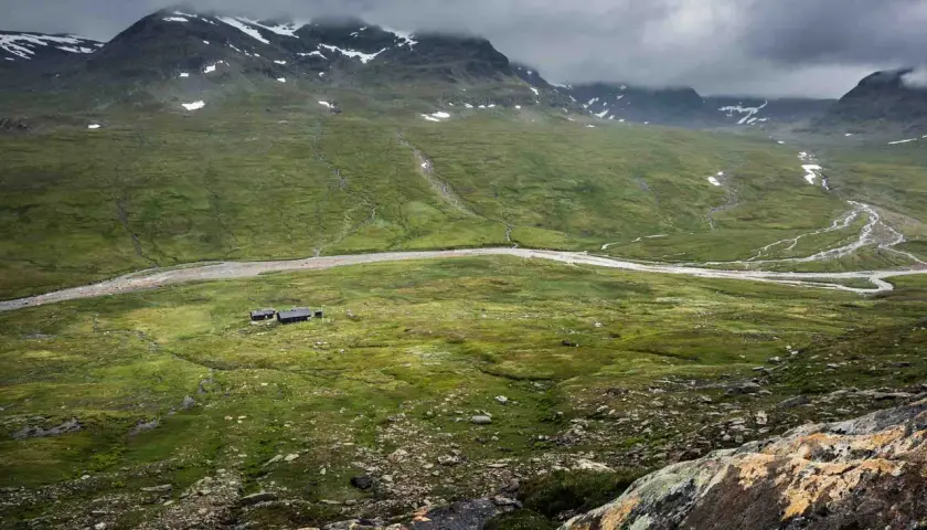 Grönskande fjällandskap med en slingrande flod och små stugor. En vy som kan vara en del av kungsleden etapper.