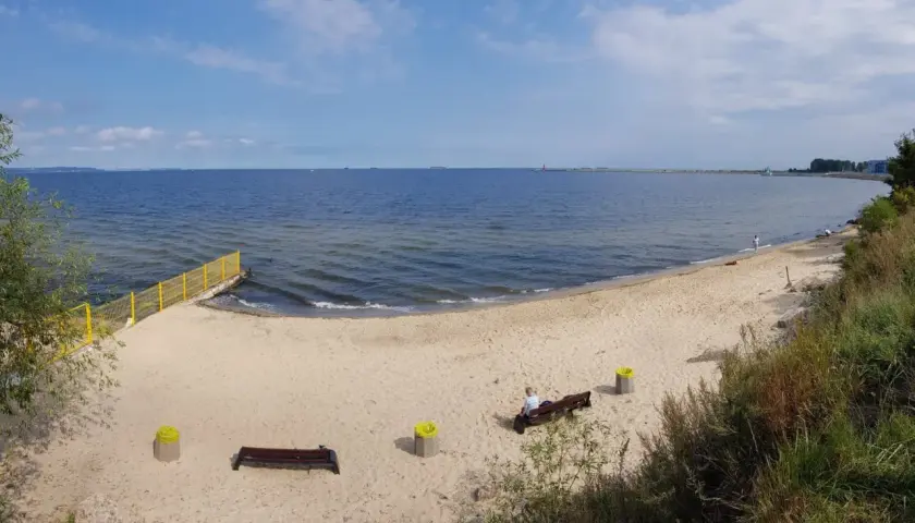 Spokojna plaża dla psów z piaskiem i morzem. Na ławce siedzi osoba, obok kosze.