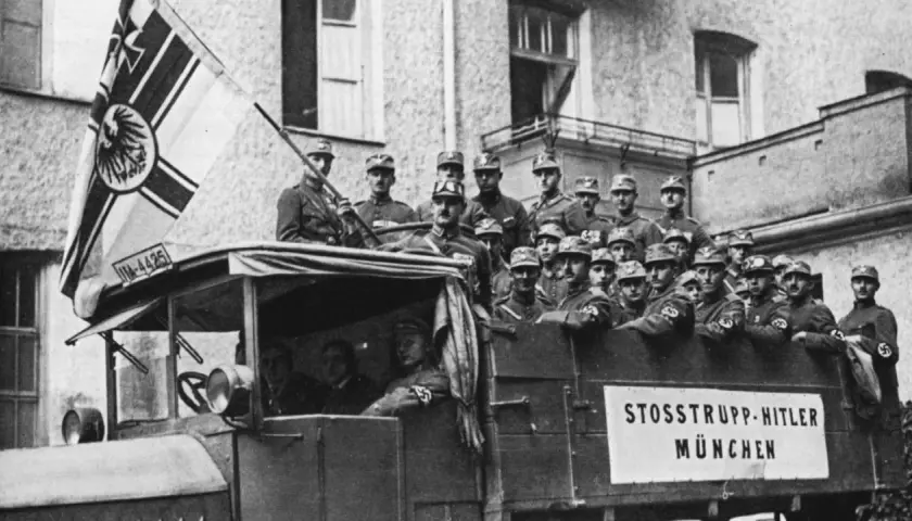 Männer in Uniformen auf einem LKW mit der Aufschrift "STOSSTRUPP HITLER MÜNCHEN". Ein Mann hält eine Fahne mit einem Adler. Dies ist ein Bild vom Hitlerputsch.
