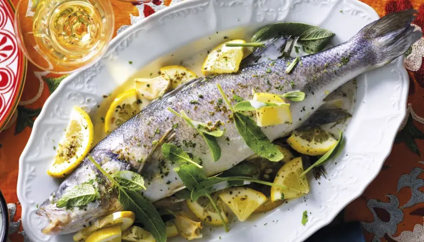 Un succulento branzino in padella, guarnito con spicchi di limone e foglie di salvia, pronto per essere gustato.