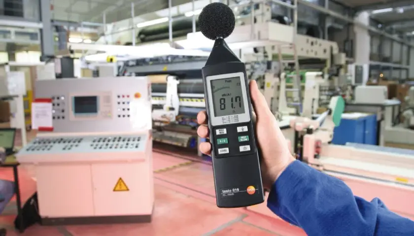 Mano sostiene sonómetro que mide 81.7 dB(A) en una fábrica, indicando el nivel de ruido.