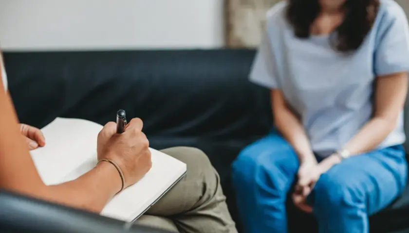 Eine Person schreibt in ein Notizbuch, während eine andere Person in blauen Jeans auf einer Couch sitzt. Dies könnte eine Szene der MDMA-Therapie sein, die bei der Heilung hilft.