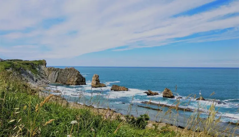 Vista de la **playa de dicido** con rocas emergiendo del mar azul y vegetación verde en primer plano bajo un cielo nublado.
