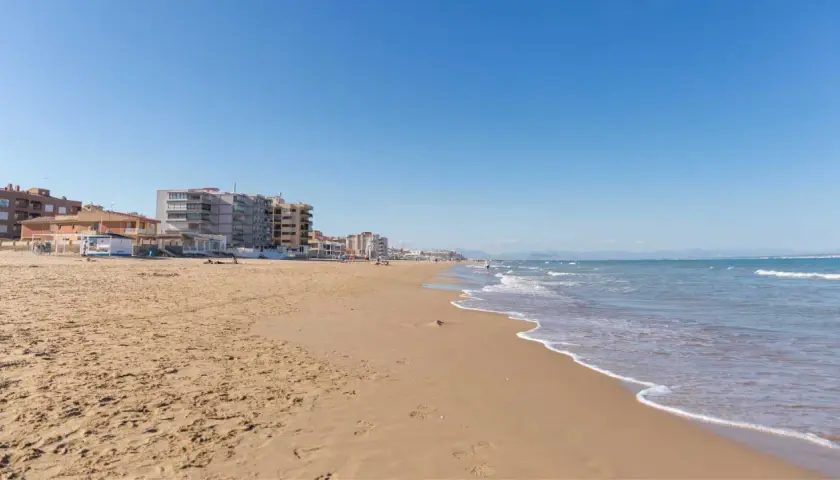 Arena dorada y mar azul en la playa La Roqueta, con edificios de apartamentos y montañas al fondo.