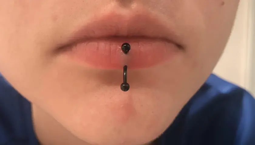 Osoba z pionowym kolczykiem w wardze (vertical labret piercing). Czarny, błyszczący kolczyk przechodzi przez środek dolnej wargi.