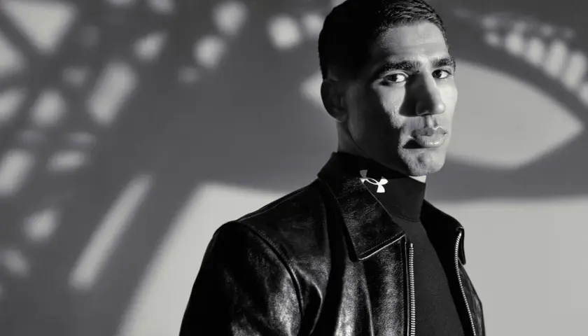 Achraf Hakimi, ein Fußballstar, blickt nachdenklich in die Kamera, trägt eine Lederjacke und ein schwarzes Rollkragenshirt mit dem Under Armour Logo.