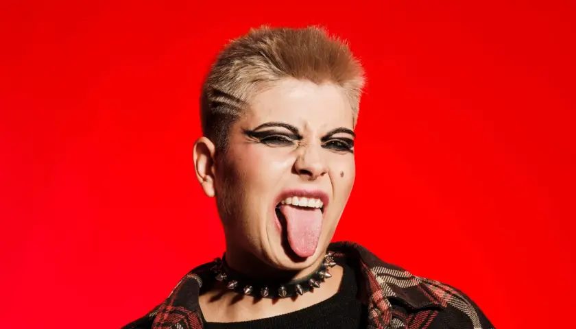 Młoda osoba z blond włosami i mocnym makijażem oczu, w kolczatce i flanelowej koszuli, wyciąga język. To kwintesencja buntu i stylu punk.