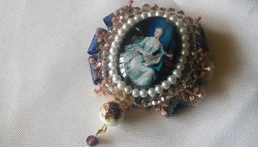Broche con camafeos, perlas y detalles florales. Una dama elegante lee un libro en un retrato ovalado, rodeada de encaje y cristales.