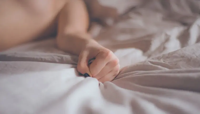 A person's hand clutches crumpled white sheets on a bed.