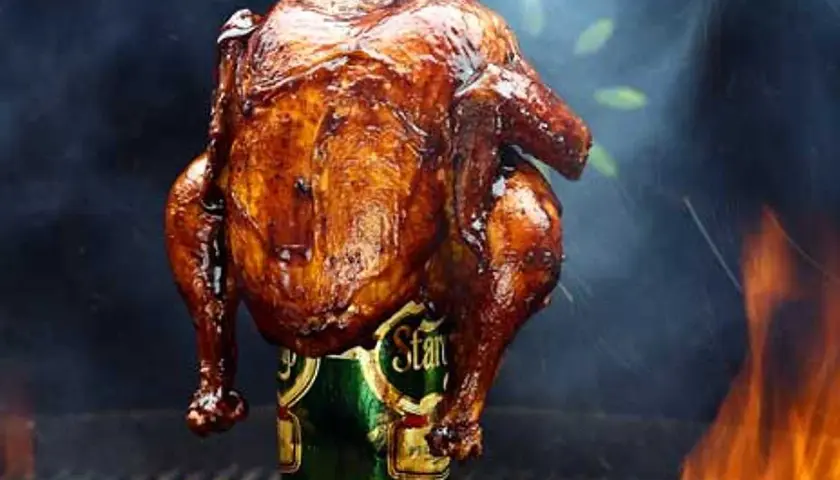 En saftig, gyllene hel kyckling tillagas stående på en ölburk, omgiven av rök och flammor. En klassisk beer can chicken.