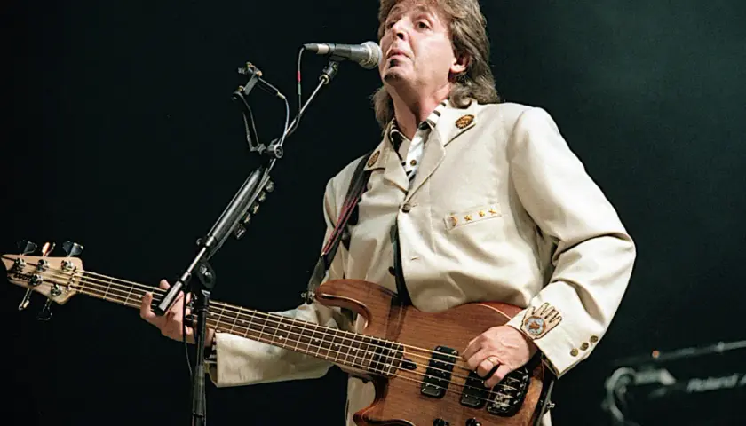 Paul McCartney z zespołu The Beatles gra na basie podczas koncertu, ubrany w elegancki beżowy garnitur.
