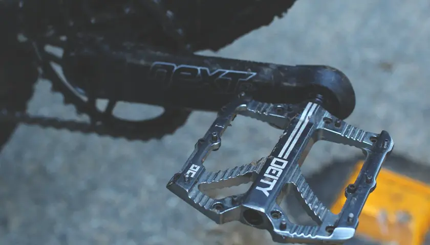 A close-up of a silver Deity pedal, ready for a Bladerunner-style descent. The crank arm reads "next SL.