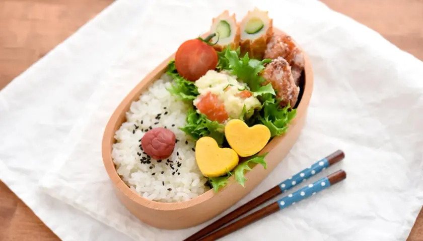 A delightful tamagoyaki bento box filled with rice, a pickled plum, potato salad, fried chicken, and cucumber rolls. Two heart-shaped tamagoyaki add a sweet touch.