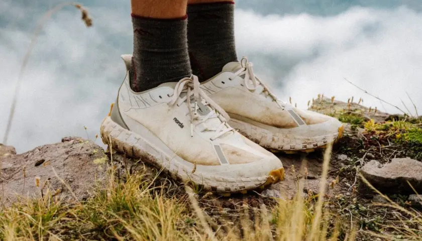 A pair of well-worn, white norda 001a trail running shoes stand on a rocky, grassy peak, ready for adventure.