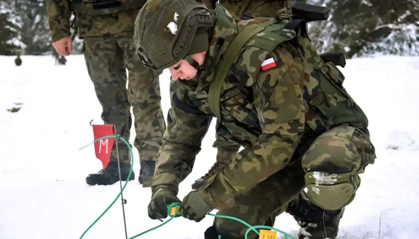 Saper w wojsku przygotowuje teren w zimowych warunkach. Żołnierz w kamuflażu pochyla się nad zielonymi przewodami, pracując ostrożnie.