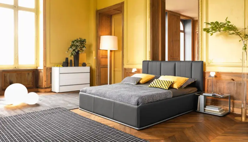 Camera da letto classica con pareti giallo ocra, letto grigio, tappeto a scacchi e arredi in legno.