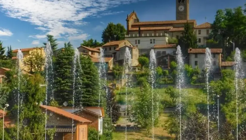 Fontane zampillanti davanti a un borgo collinare con chiesa e campanile. Un'immagine che invita a scoprire cosa vedere a Fortunago.