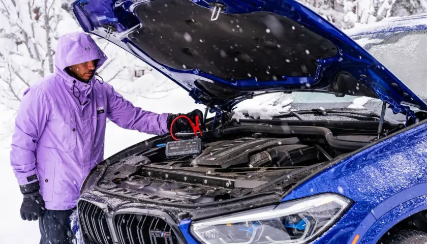 Mężczyzna w fioletowej kurtce podłącza booster do akumulatora niebieskiego BMW. To rozwiązanie, co może pobierać prąd w samochodzie na postoju, gdy jest zimno.