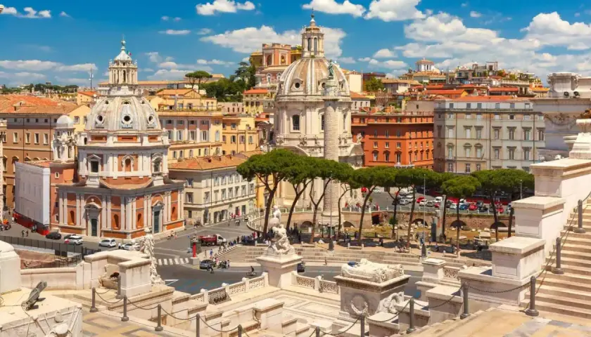 Vista panoramica di Roma con l'Altare della Patria in primo piano, cupole imponenti e palazzi storici sotto un cielo azzurro con nuvole bianche.