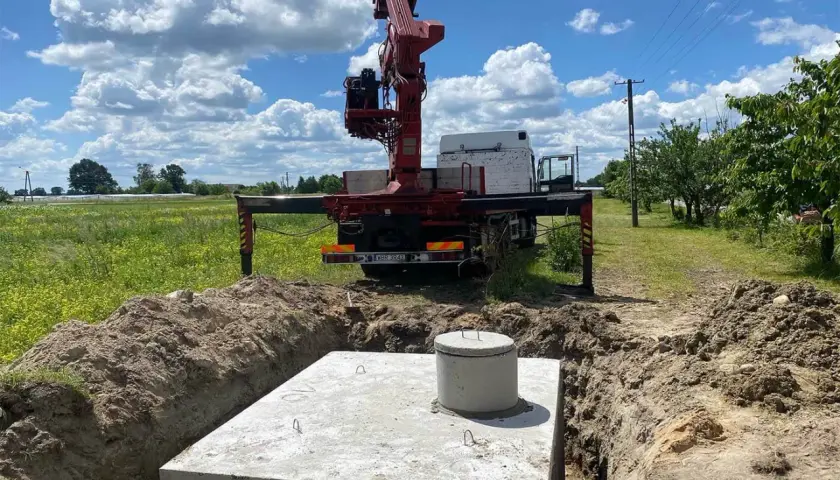 Montaż szamba: betonowa płyta i kominek wystają z wykopu, obok stoi ciężarówka z dźwigiem.