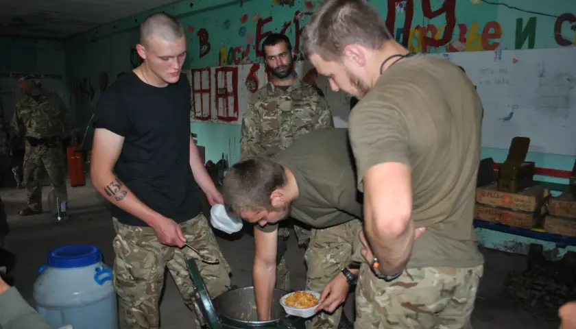 Żołnierze w mundurach kamuflażowych przygotowują posiłek. Jeden z nich, z tatuażem "13" na ramieniu, nabiera jedzenie łyżką. W tle widać napisy na ścianie, sugerujące przygotowania do szturmu.