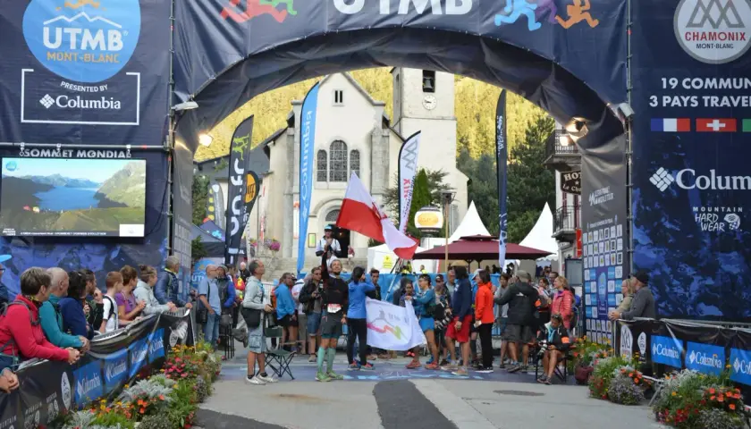 Uczestnicy Ultra-Trail du Mont-Blanc świętują pod łukiem startowym. W tle kościół i tłum widzów.