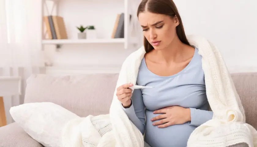 Donna incinta preoccupata guarda il termometro, avvolta in una coperta. La febbre in gravidanza è un sintomo da non sottovalutare.